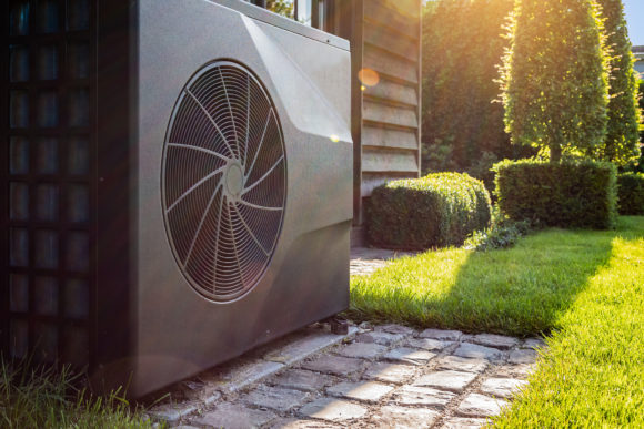 Close-up of black full inverter heat pump outside in the garden, near wooden pool house on a sunny day. Lens flare on the image.